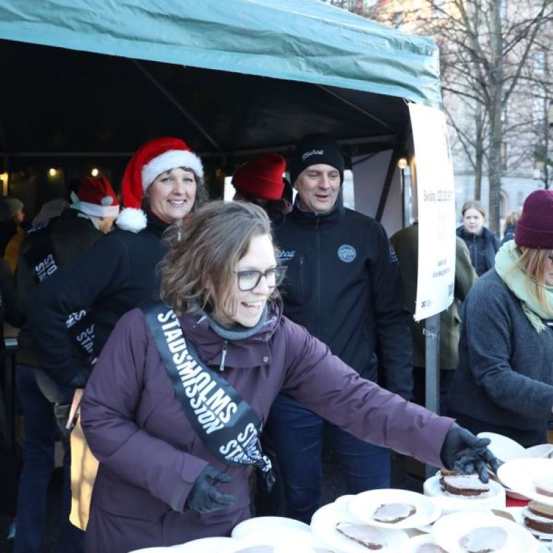 Kvinna står framför ett tält i Kungsträdgården och serverar smörgåsar med julskinka. Över bröstet har hon ett band där det står Stockholms Stadsmission. I bakgrunden två personer som ler. 