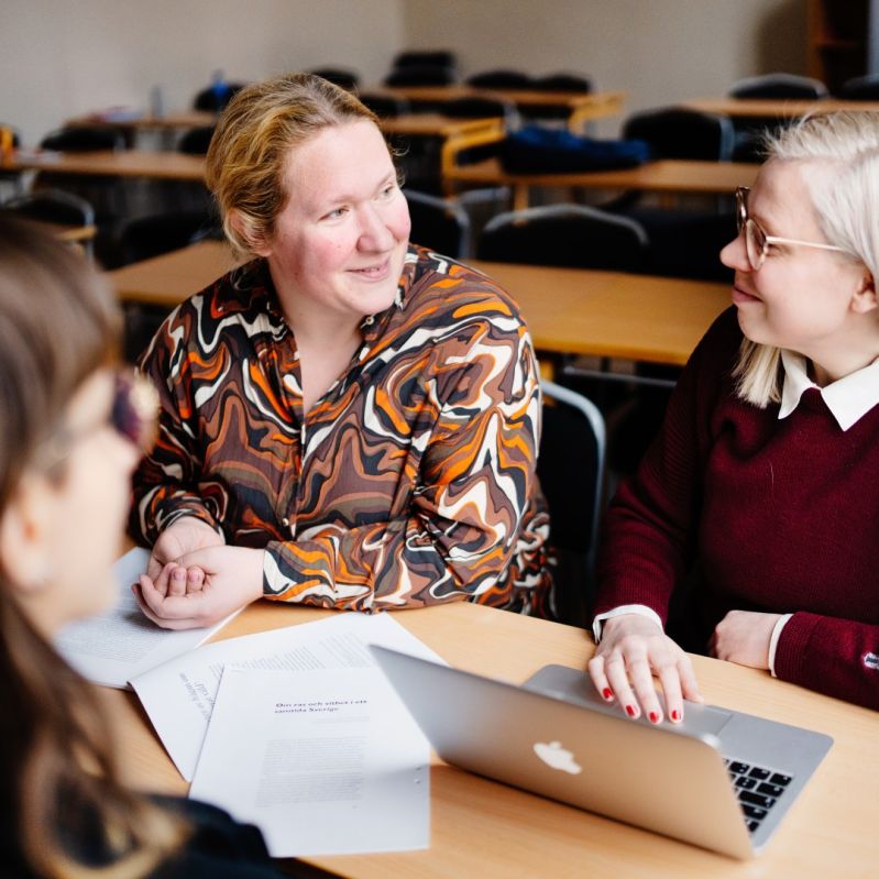 Tre kvinnor sitter runt ett bord och diskuterar i ett klassrum.